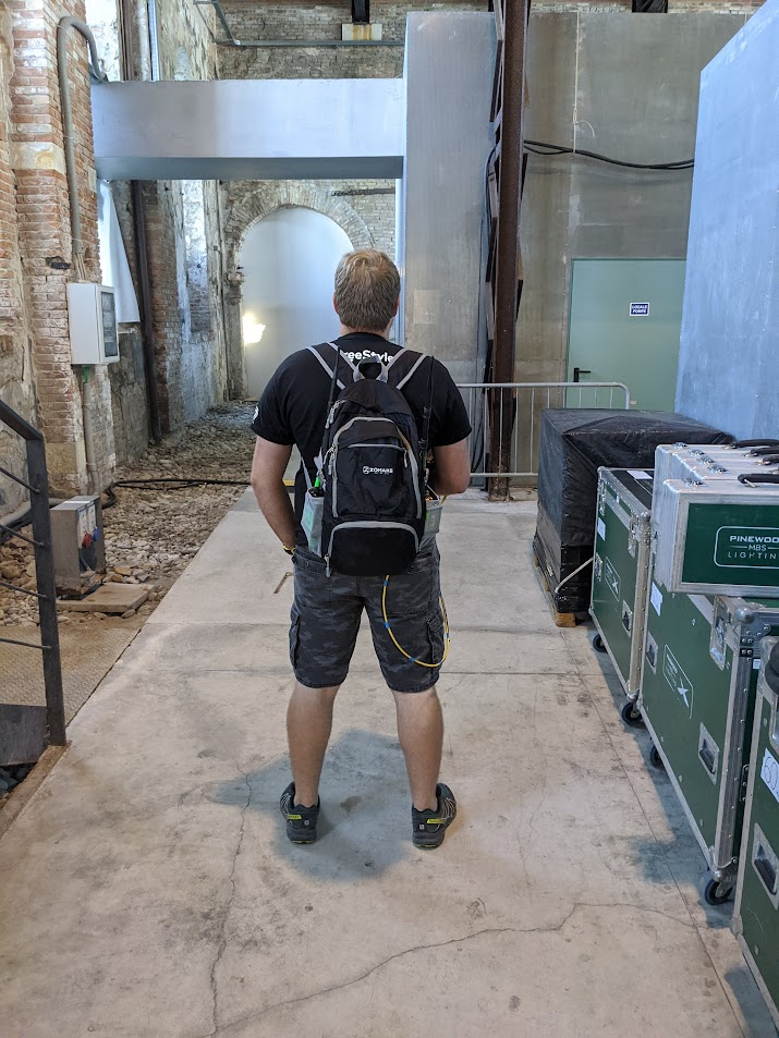 Man with backpack explores rustic hallway, surrounded by equipment cases, inside an industrial-style building.